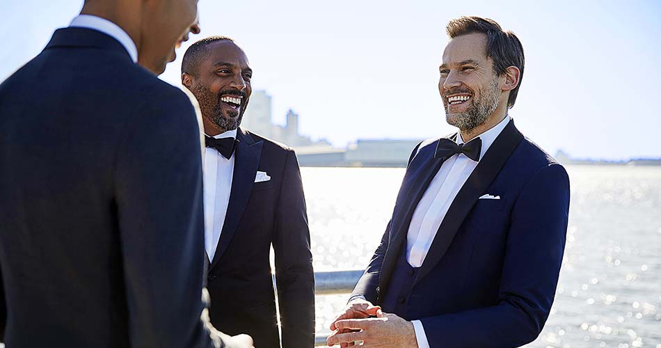 three men wearing blue tuxedo laughing against river backdrop 