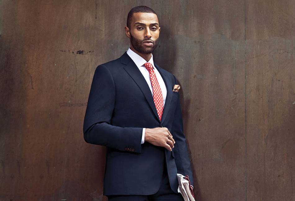 man wearing navy suit with orange polka dot tie and orange pocket square leaning against a dark wooden wall