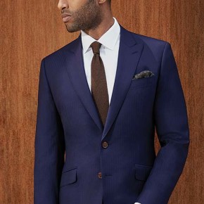 man wearing a navy suit with brown knitted tie against a wooden wall