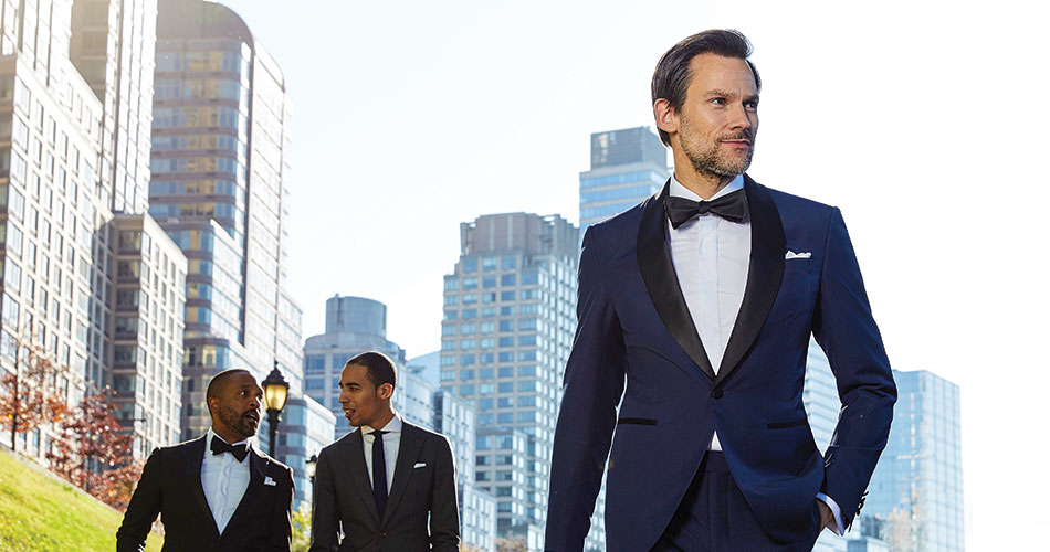 man walking outdoors wearing blue tuxedo with two other men wearing a black tuxedo and a charcoal suit behind him