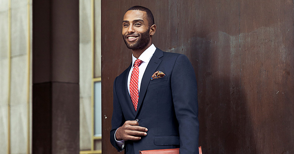 man in navy suit and dotted orange tie standing against a wooden wall