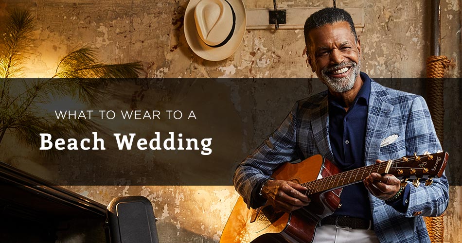man wearing blue checked blazer holding a guitar with text overlay "what to wear to a beach wedding"